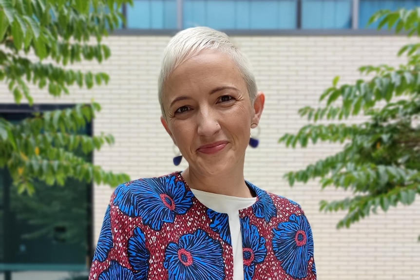 A short-haired woman leaning on a table while smiling to the camera.