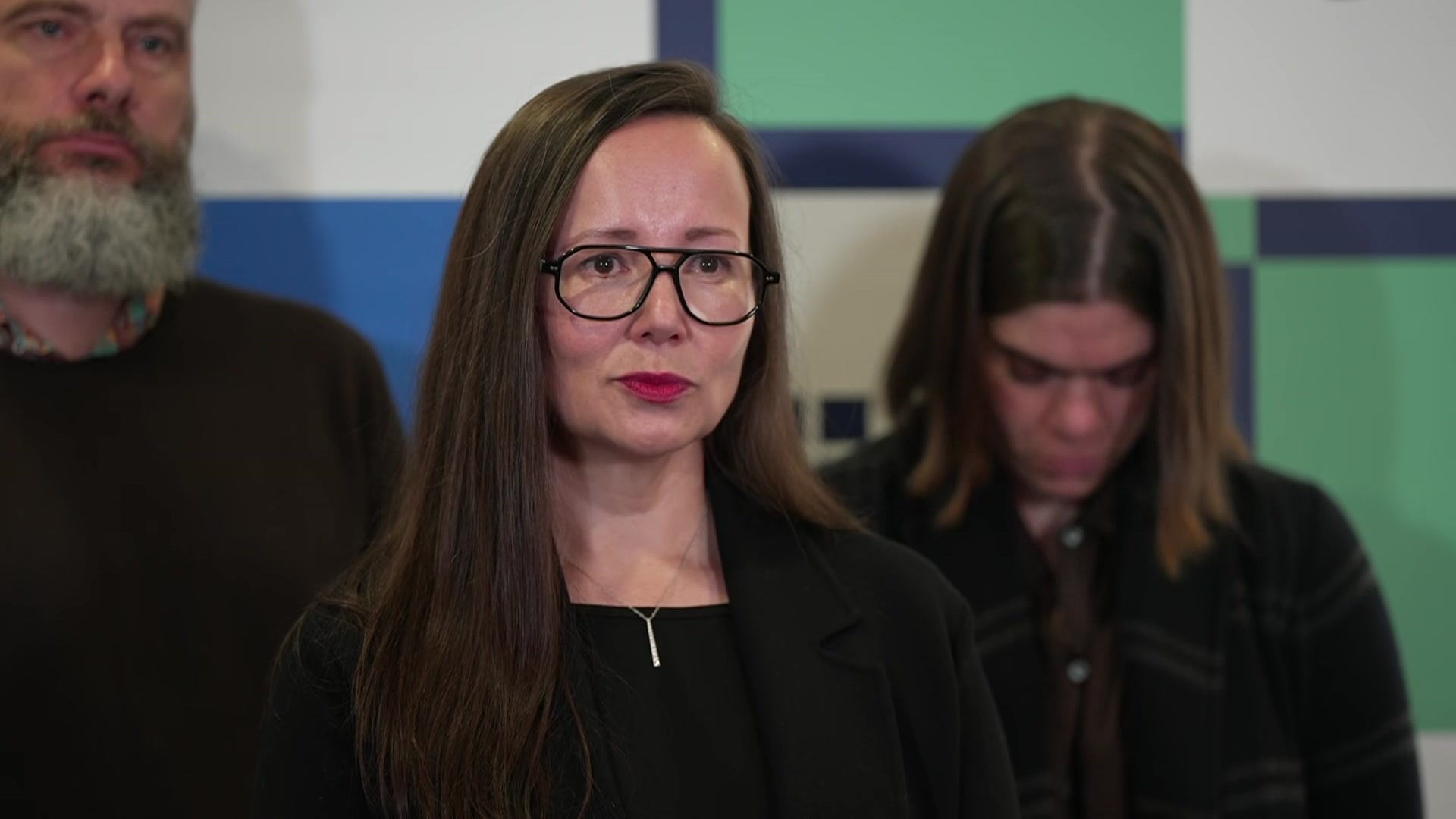 A woman with long dark hair, dark rimmed glasses and a black top stands in front of two people staring off camera.