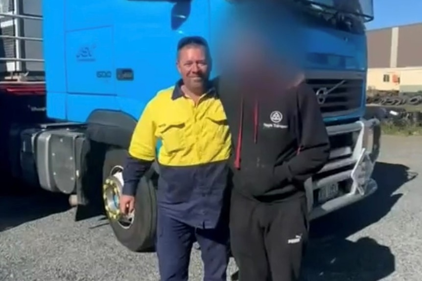A man wearing a yellow and navy workshirt, standing in front of a truck next to another person, whose face is blacked out.