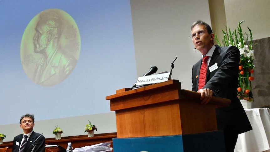 A man stands  at a podium with Thomas Perlmann written on a name plate. A noble prize logo is projected onto a nearby screen.