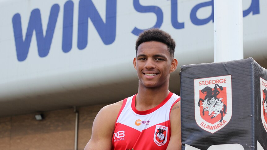 A smiling young man leans against a football post wearing a sleeveless red and white jersey.