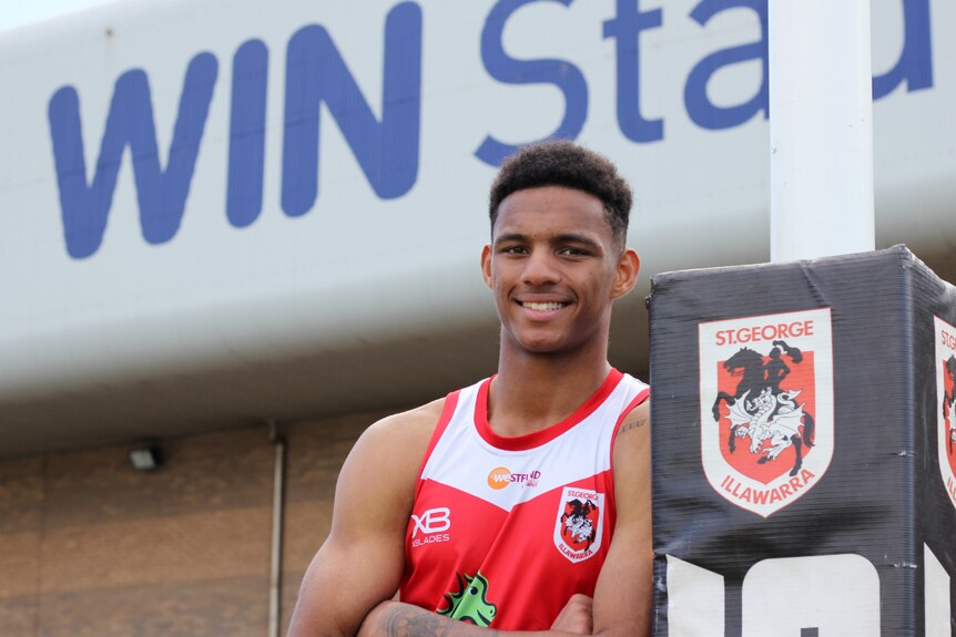 A smiling young man leans against a football post wearing a sleeveless red and white jersey.