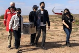 People wearing black face masks carrying a metal box with gauges on it in the desert