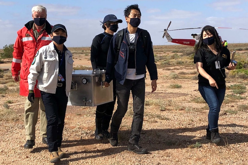 People wearing black face masks carrying a metal box with gauges on it in the desert