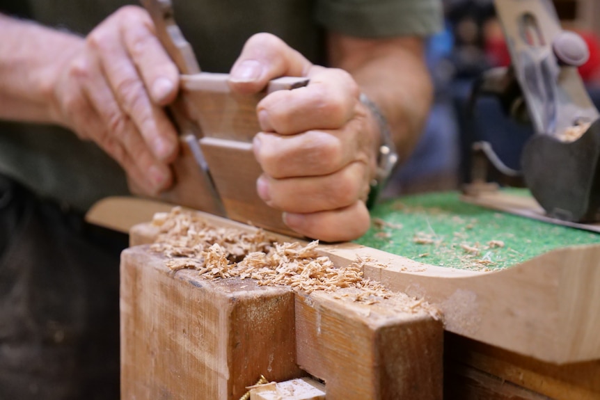 A man's hands holding onto a woodworking tool, with wood shavings next to a piece of timber.