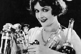 A 1920s black and white photo of a woman holding many bottles of alcohol