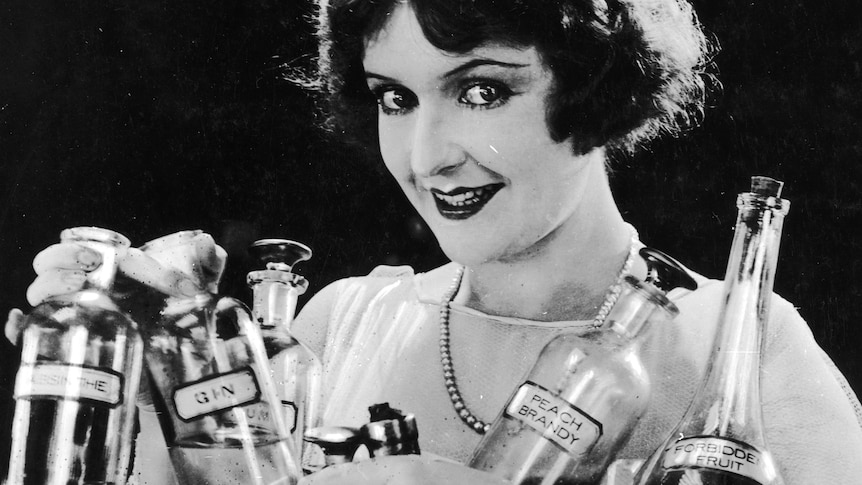 A 1920s black and white photo of a woman holding many bottles of alcohol