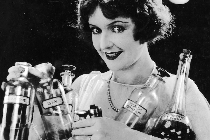 A 1920s black and white photo of a woman holding many bottles of alcohol
