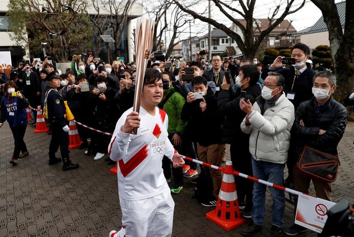 A man wearing a white tracksuit with a red stripe across the top runs while holding the Olympic torch with people watching.