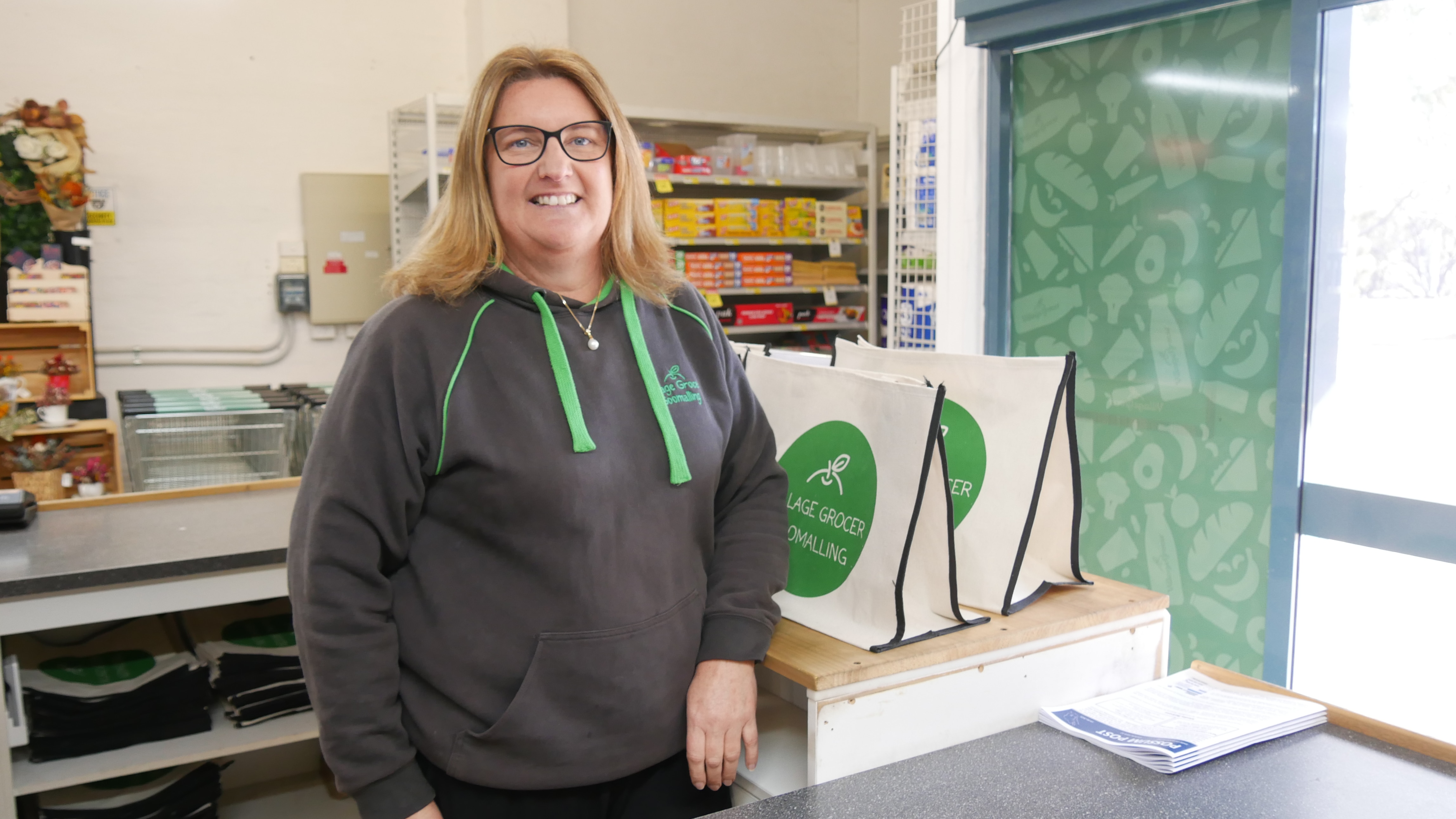 A woman in a grey and green hooded jumper stands in a grocery shop smiling at the camera.