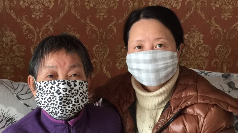 A woman wearing a facemask leans over an elderly woman laying down and wearing a face mask.