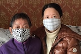 A woman wearing a facemask leans over an elderly woman laying down and wearing a face mask.