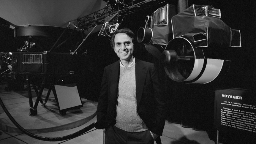 Black and white photo of astronomer Carl Sagan standing in front of equipment.