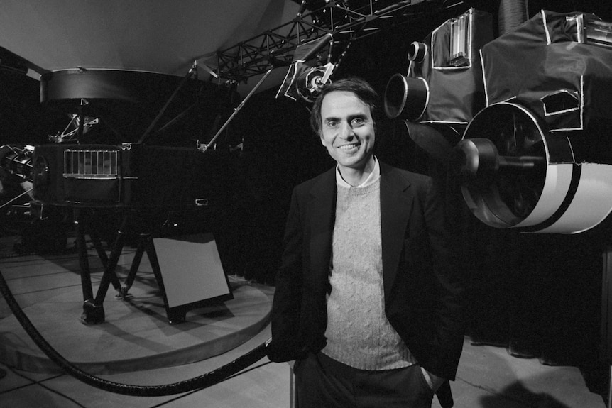 Black and white photo of astronomer Carl Sagan standing in front of equipment.