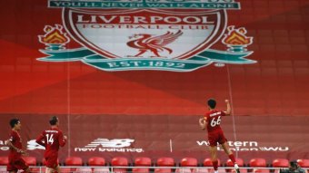A Liverpool player leaps in the air after scoring, with a big Liverpool banner in the background.