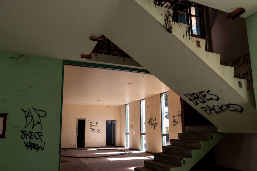 A dilapidated staircase, covered in graffiti can be seen, the room behind it is gutted, the windows have no glass.