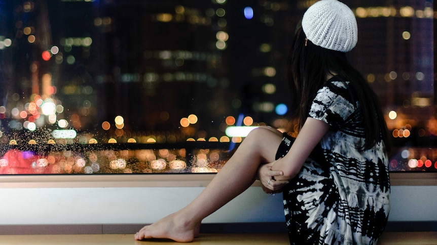 girl sits on balcony alone, looking longingly outside at the city lights