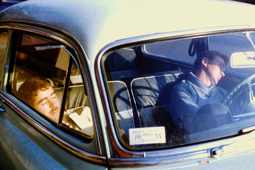 Two men slouch and sleep in the front seats of a car in 1955