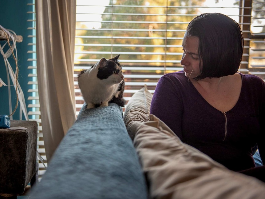 A dark-haired woman sits on a couch and looks at a cat.