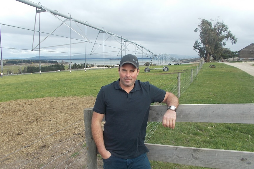 A man leans on a fence in front of a large irrigation pivot at a farm.