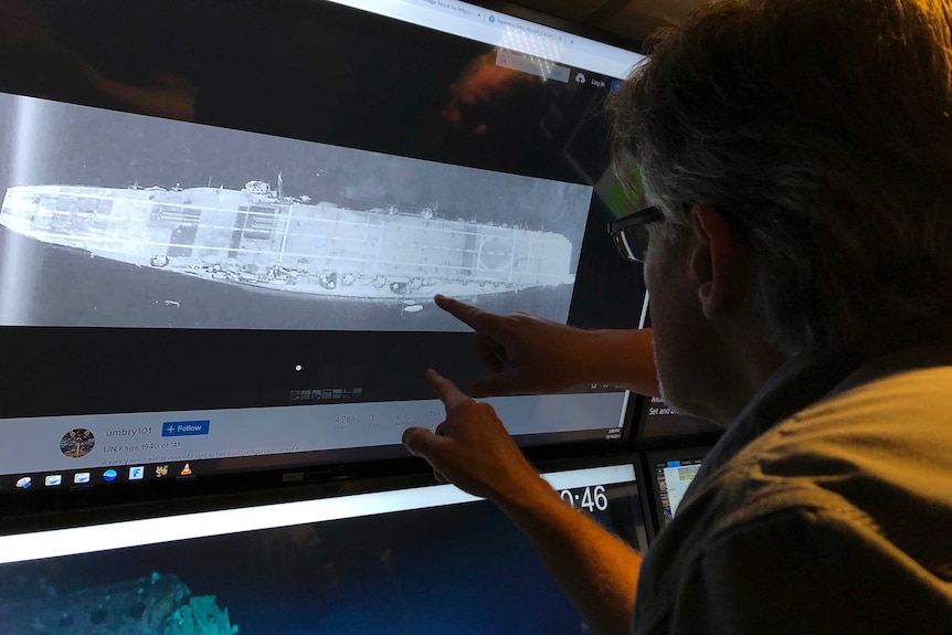 A man is looking at a warship that is underwater on a large screen and pointing at it