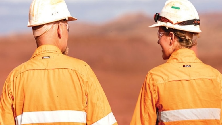 One male and one female mining worker wearing orange high vis stand with their backs to the camera