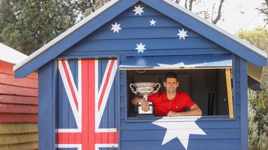 Novak Djokovic stands in a cabin painted in the Australian flag as he holds the trophy after winning the Australian Open. 