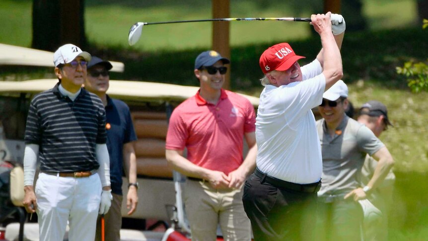 Trump swings golf club wearing red cap with USA on it while Abe and others look on