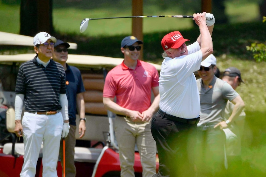 Trump swings golf club wearing red cap with USA on it while Abe and others look on