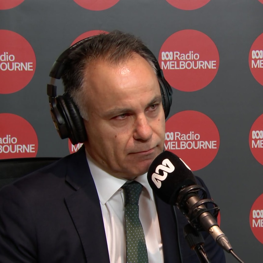 A man in a suit with a green tie sits in front of a mic with ABC Radio Melbourne logos in the background
