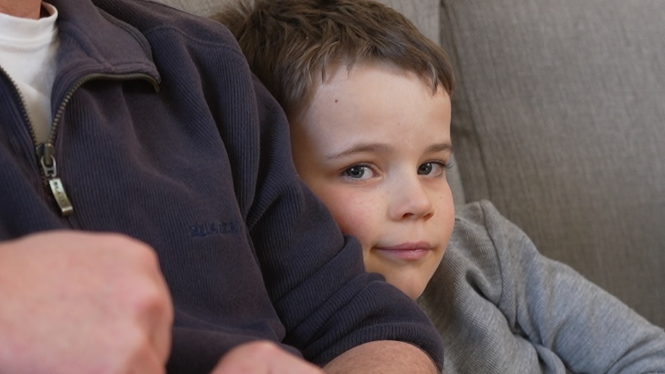 Rohan smiles, sitting next to an adult on the couch. He has brown hair.