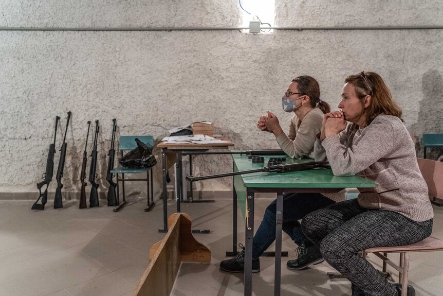Two women sit on chairs at a table with guns on it. More guns are against a wall in the background.