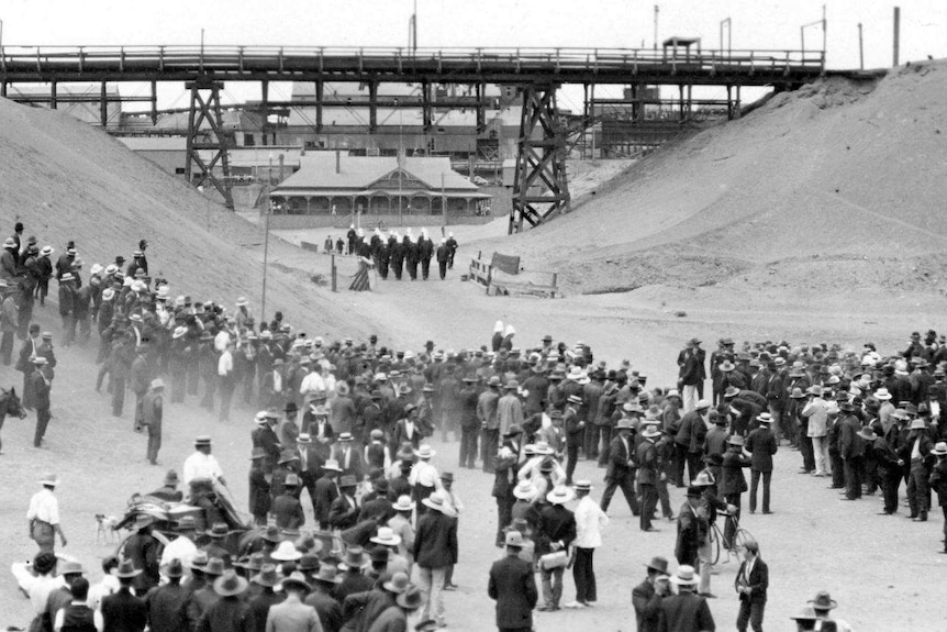 A black and white archive photo from 1909 showing a crowd of people standing at a mine site. Row of police leave the site