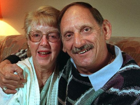 A heterosexual couple in their seventies, a blonde woman with glasses and a man wearing a patterned jumper, embrace and smile.