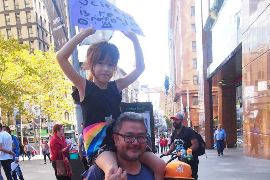 A young girl holds a sign sitting on her fathers shoulders while a young boy stands next to the father holding a sign