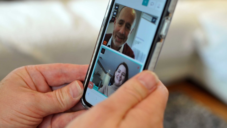 Hands hold an iPhone. On the screen is a video call with windows showing a man talking to a woman.