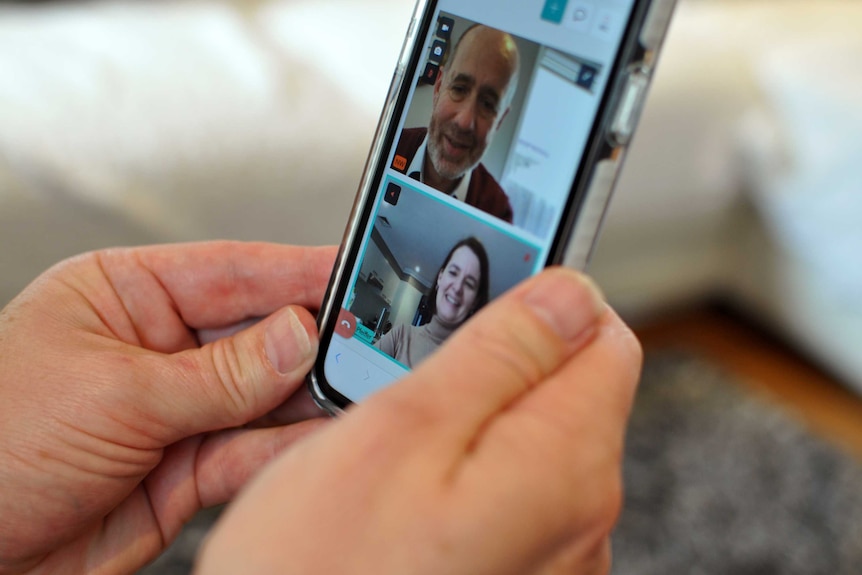 Hands hold an iPhone. On the screen is a video call with windows showing a man talking to a woman.