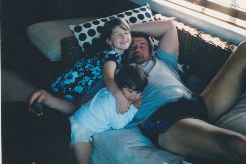 A man holds out a television remote while laying on a couch and two little girls hug him.