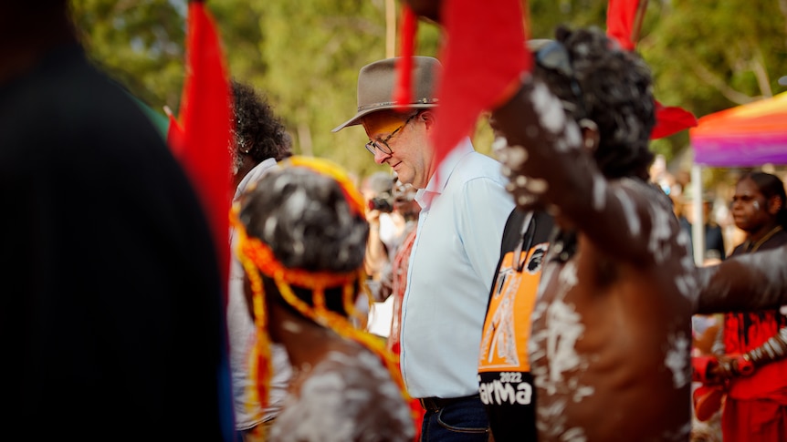 Albanese in a brimmed hat looks down, surrounded by young Indigenous boys in body paint, dancing