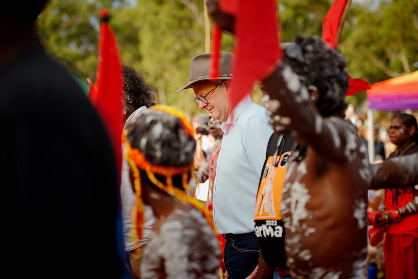 Albanese in a brimmed hat looks down, surrounded by young Indigenous boys in body paint, dancing