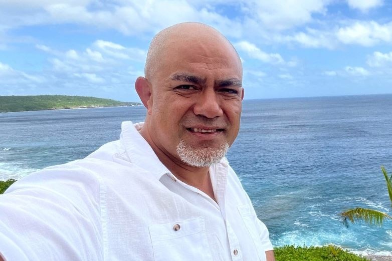 Man with bald head in white shirt takes a selfie standing infront of ocean. 