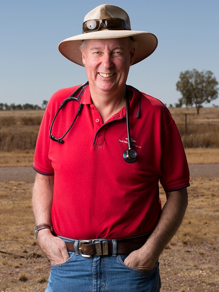 Man wearing wide-brimmed hat and red polo shirt with stethoscope around his neck in bush setting.