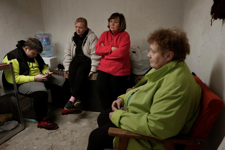A group of women stand around inside a bomb shelter with sombre expressions. 