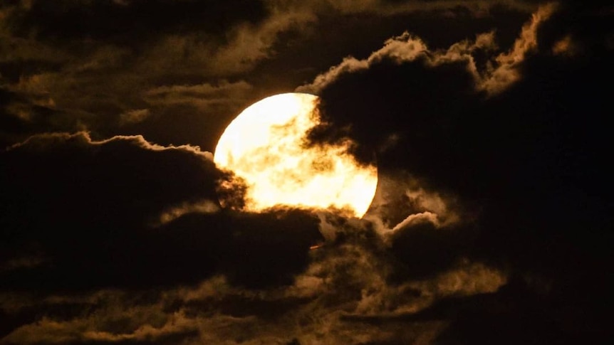 A bright glowing moon behind dark clouds at night time. 
