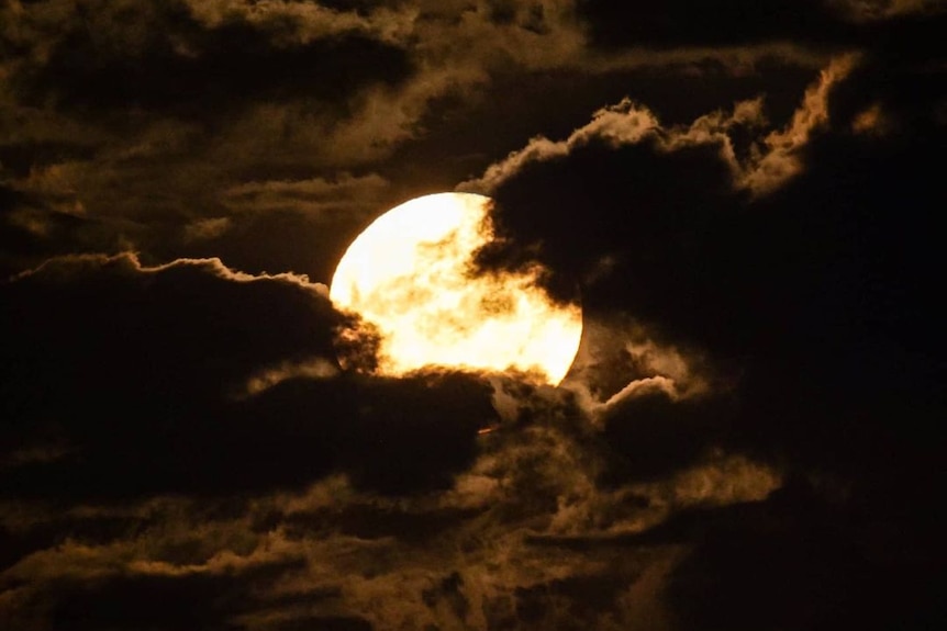 A bright glowing moon behind dark clouds at night time. 