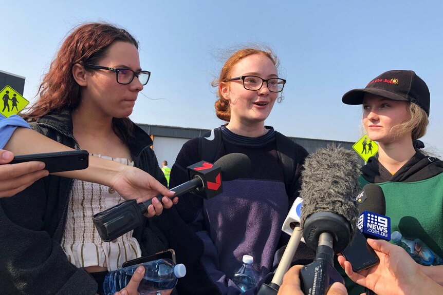 Three teenage girls speak to reporters holding media-branded microphones.