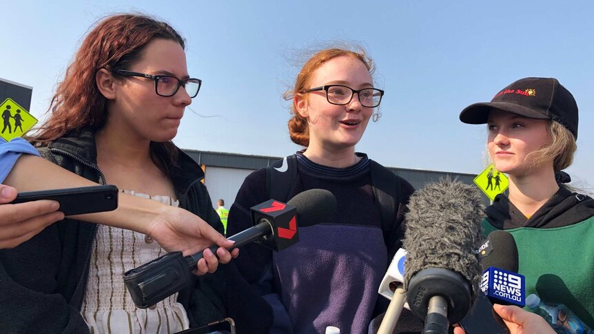 Three teenage girls speak to reporters holding media-branded microphones.