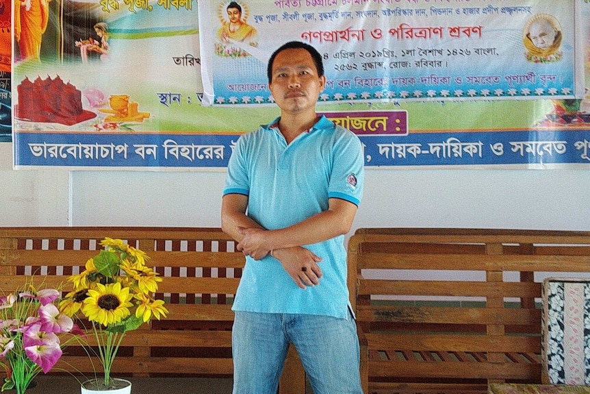 A man with black hair and a blue shirt stands in front of Bangladeshi posters.