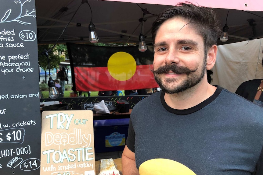 Tim Sculthorpe stands at a food stall smiling. An Aboriginal flag is in the background.