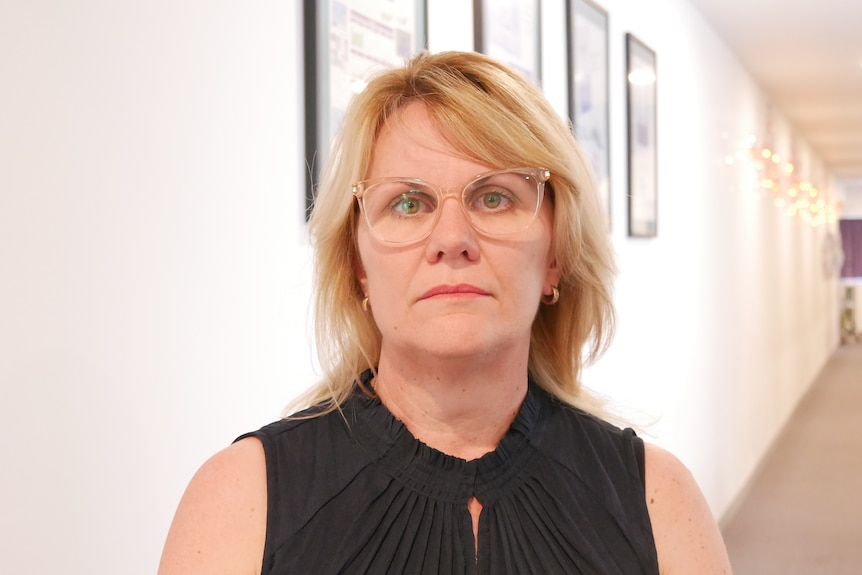 A blonde woman looks at camera behind her is a hallway with photo frames lining the wall.